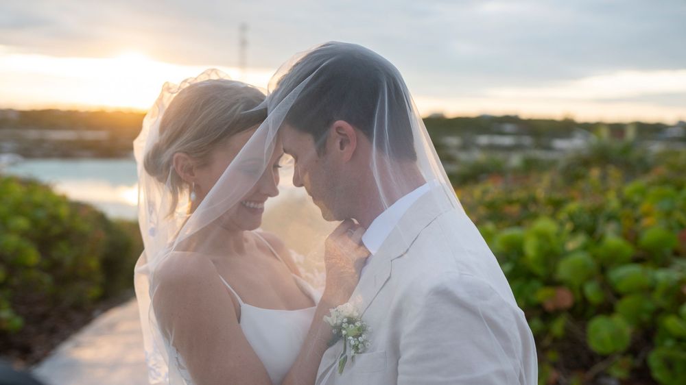 Romantic newlywed couple shares intimate moment at sunset with glowing backlight through bridal veil.