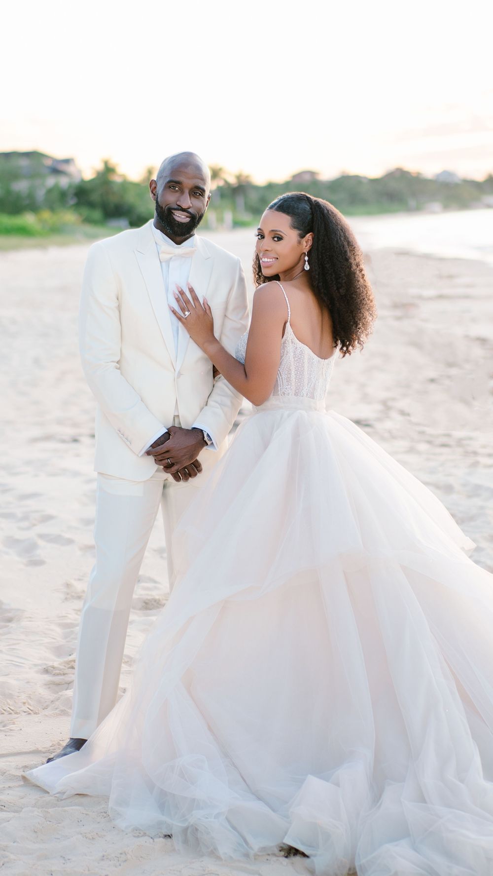 A glamorous beach wedding photo of a couple in all white attire posing on the sand.