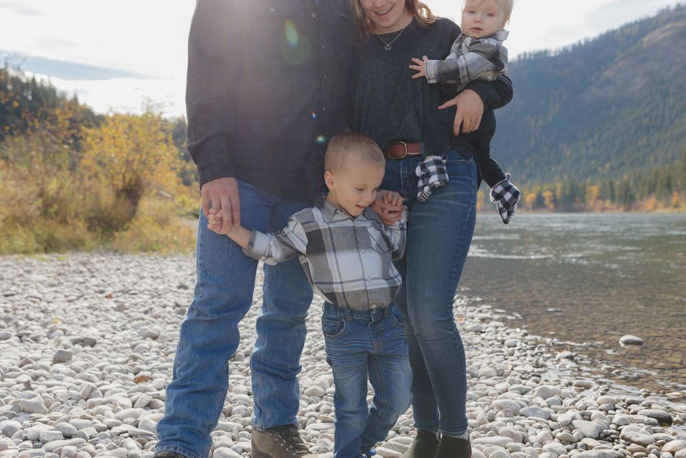 A scenic family moment on a rocky riverbank with mountains and autumn trees in the background.