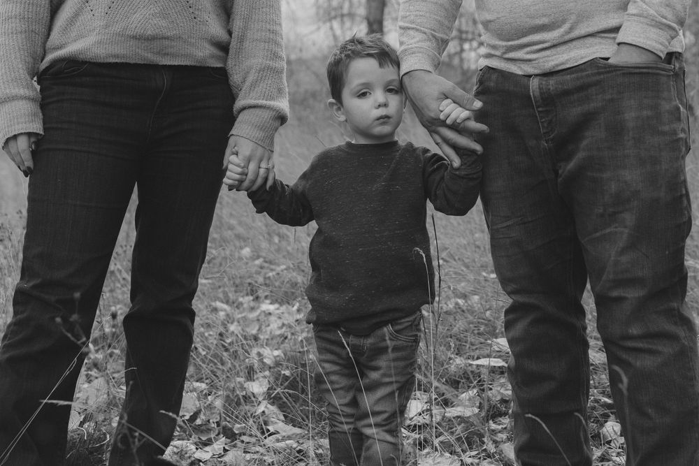 Black and white photo showing small child in dark sweater holding hands with two adults in jeans outdoors.