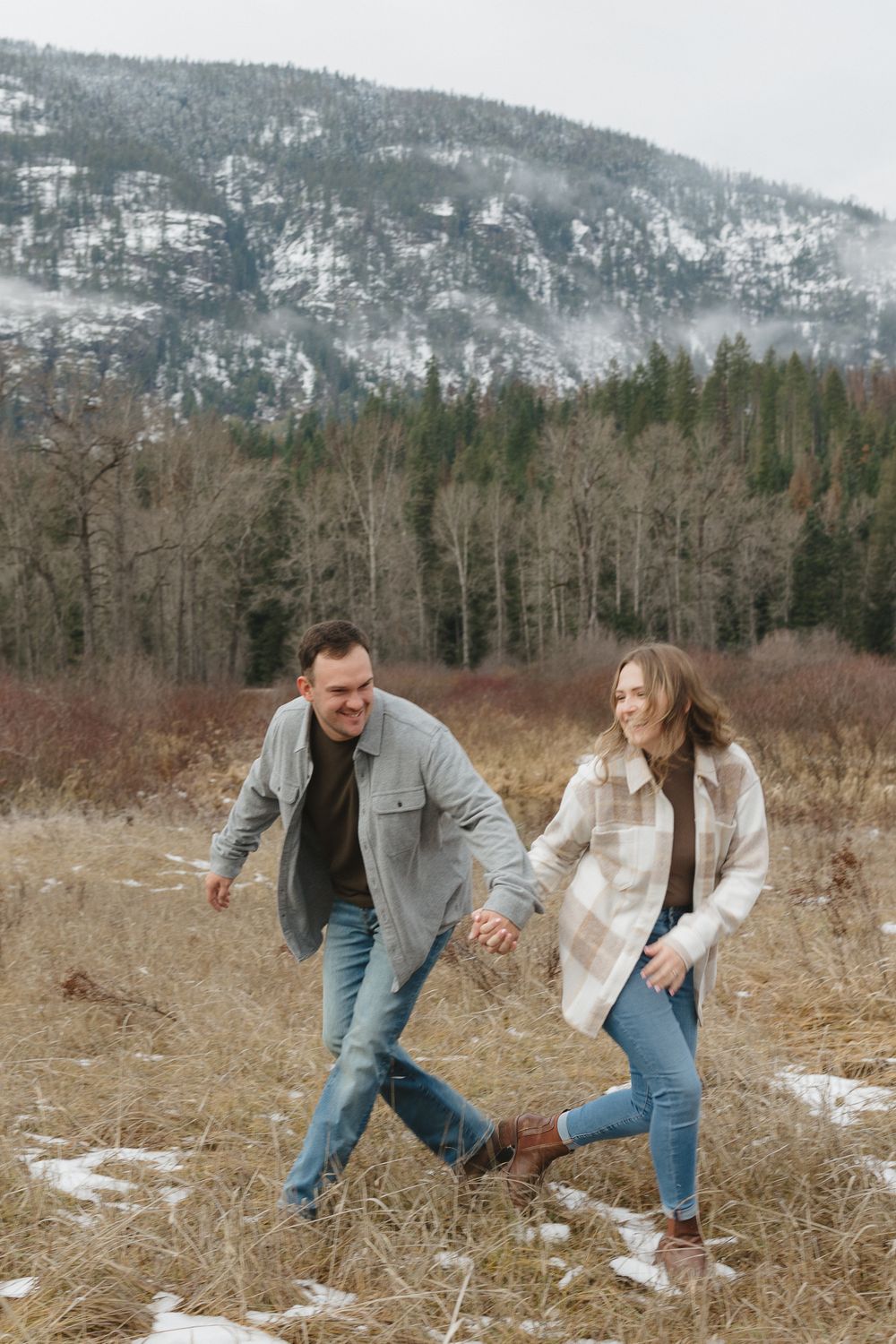 A couple holds hands while running through a snowy field with evergreen mountains in the background on a winter day.