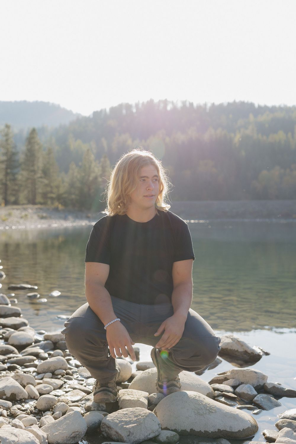 A person in a black t-shirt crouches on rocky riverbank with sunlit forest and turquoise water in background.