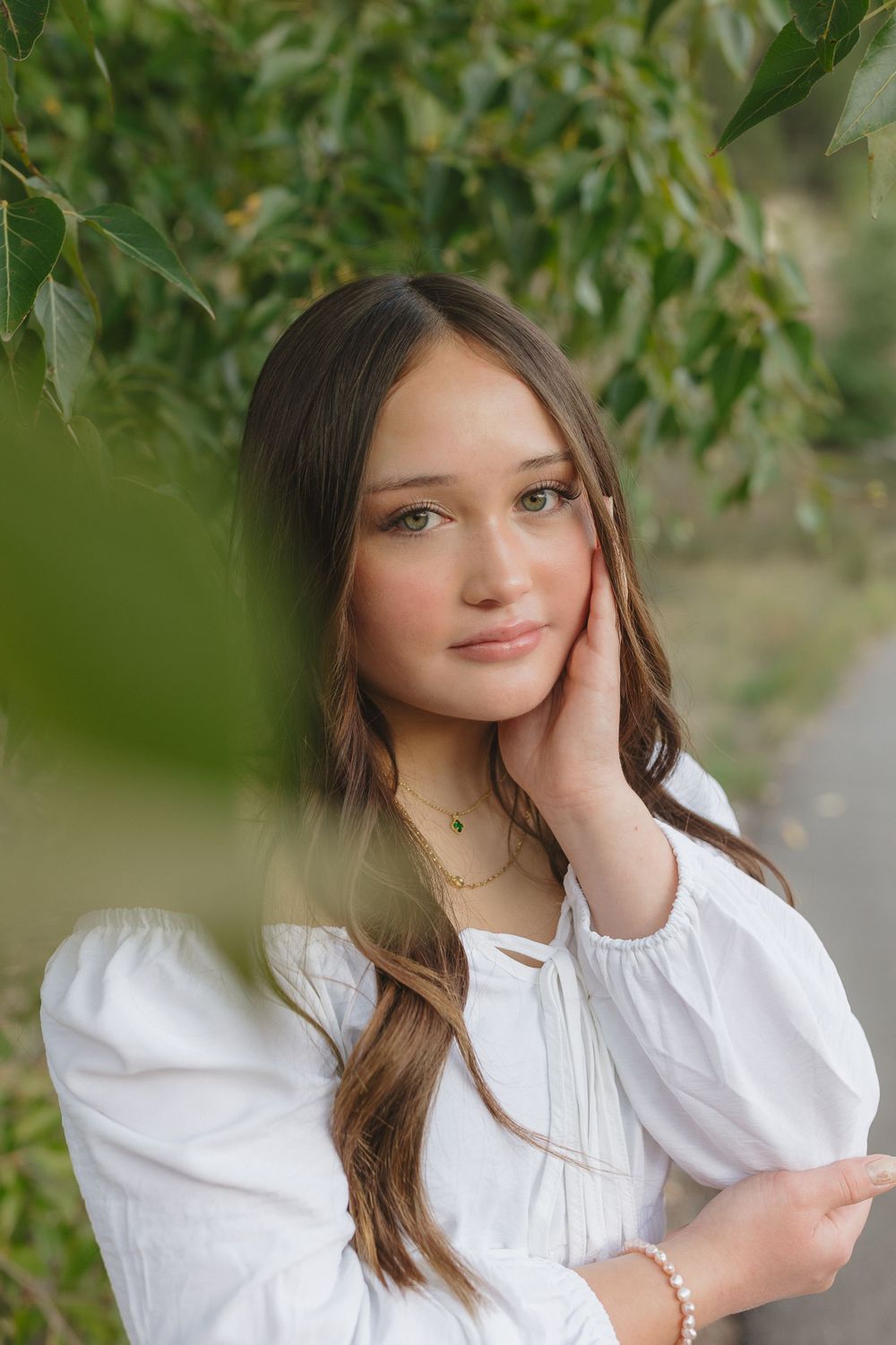 Gentle outdoor portrait with soft natural lighting through green foliage, featuring white peasant top and delicate jewelry.