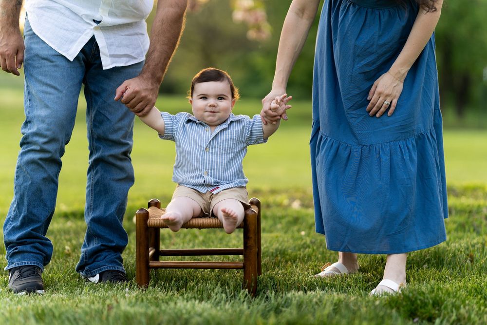 Small child sits on a wooden bench while being supported outdoors in a grassy setting between two adults.