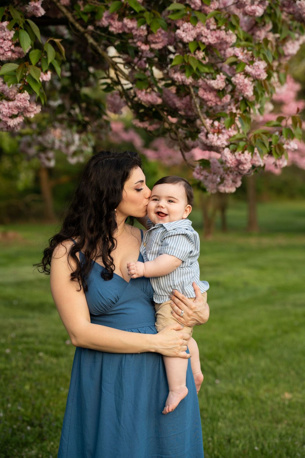Mother in blue dress holds and kisses happy baby under blooming pink cherry blossoms in a spring garden.