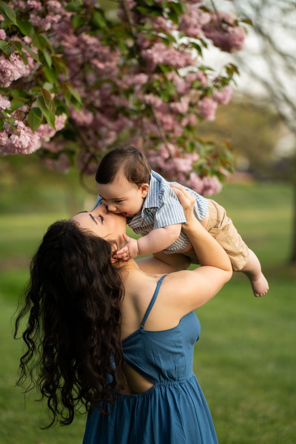 A tender moment captured under pink cherry blossoms as a baby is lifted up in a blue dress.