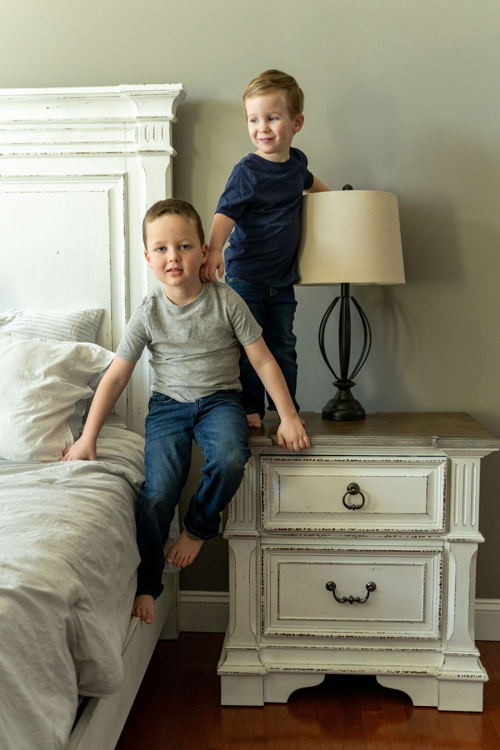 Two children playing together on white vintage furniture in a bedroom with a table lamp beside them.