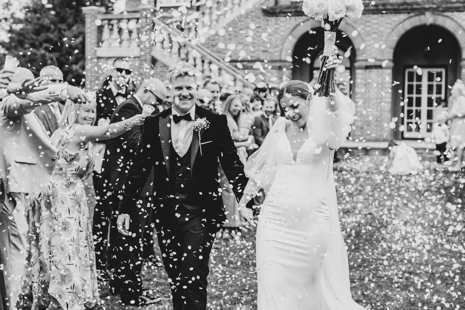 Black and white photo of a bride and groom walking through confetti shower surrounded by cheering guests at elegant venue.