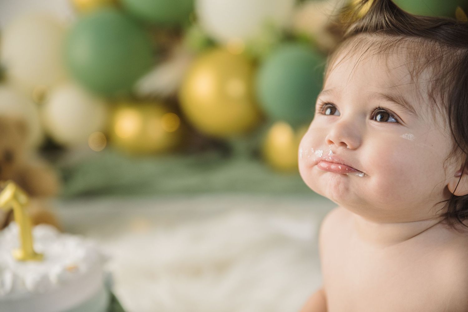 Baby smiling at first birthday celebration with cake and gold and green balloon decorations.