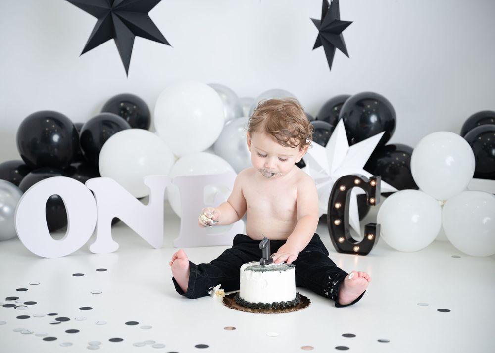 Baby in black and white themed cake smash photo session with balloons and confetti on white backdrop.