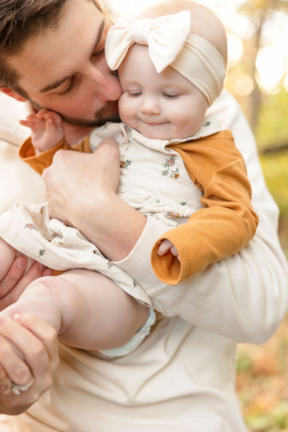 Sweet tender moments captured in autumn sunlight as parent shares loving kisses with baby wearing white and gold.