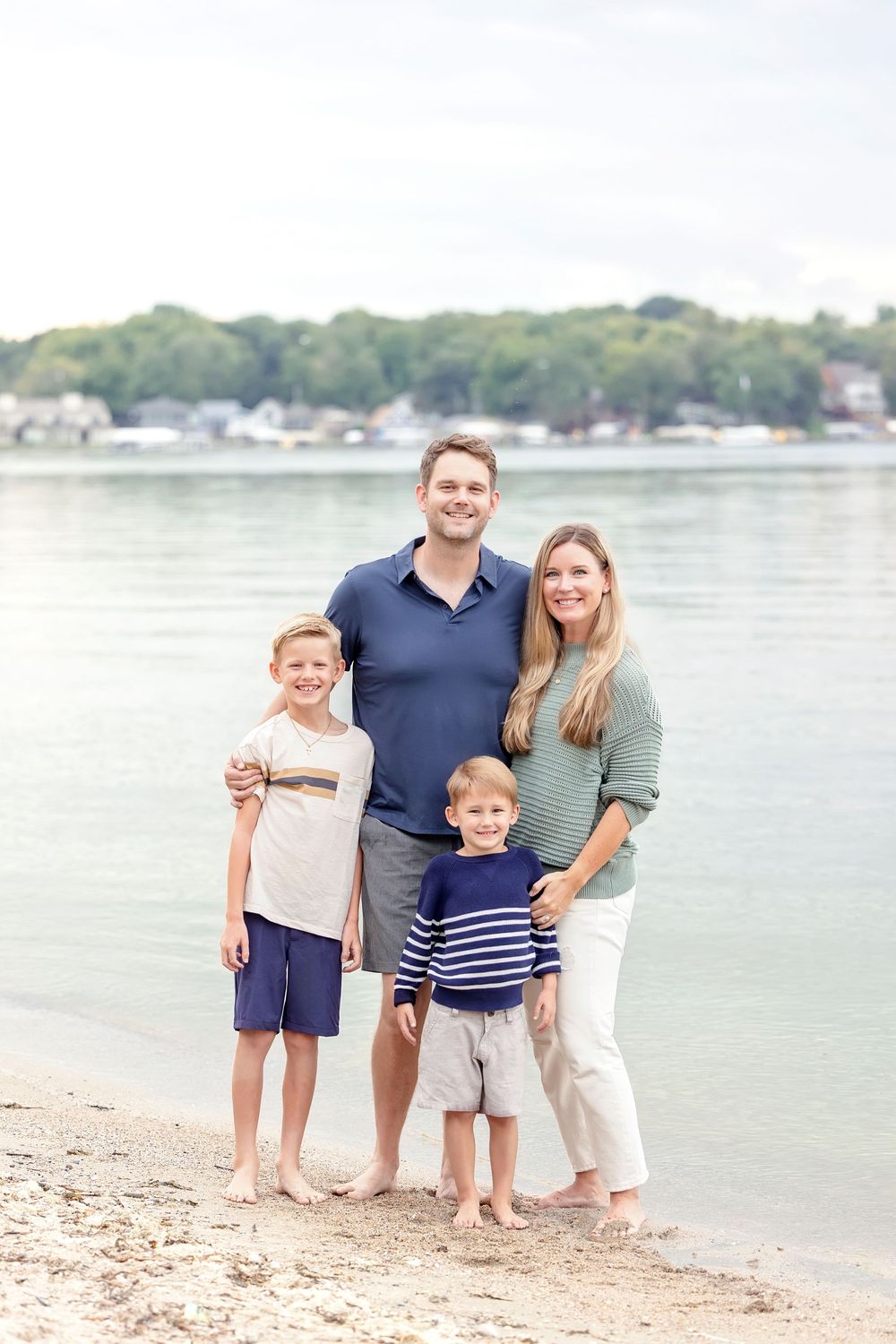 Family of four standing together for a portrait on a beach with water in background.
