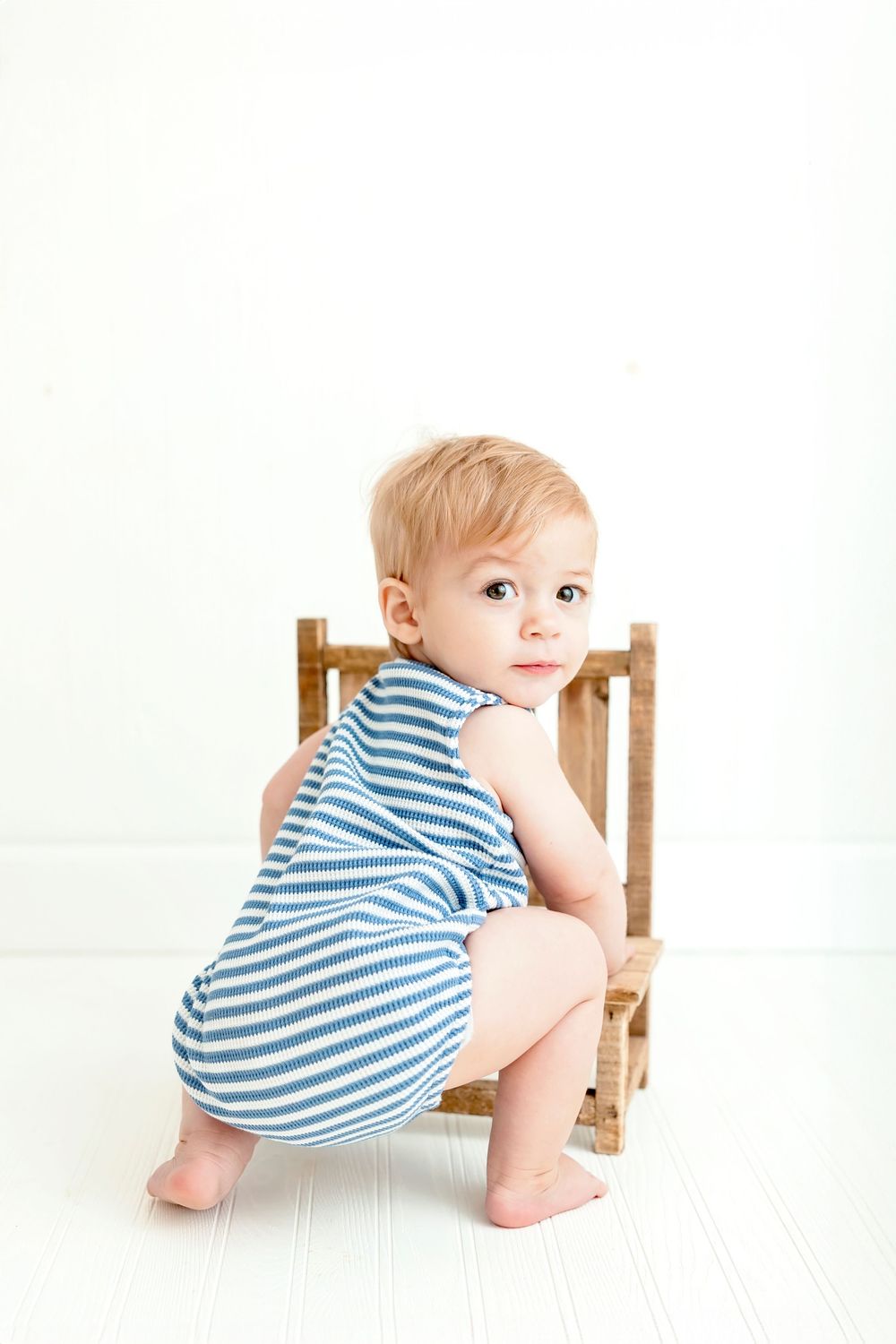 Baby in blue striped romper crawls and explores wooden chair during photo session.