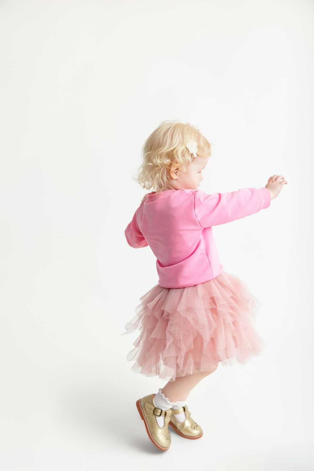 Toddler girl in pink hoodie, tutu skirt, and gold shoes dancing against a white background.