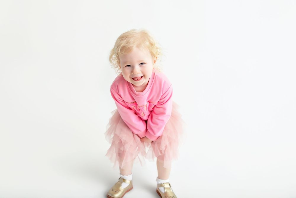Joyful toddler girl in pink tutu skirt and sweater laughing and leaning forward on white background.