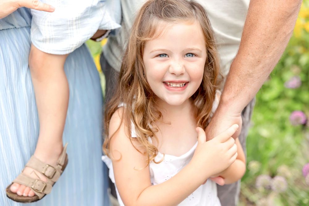 A young child in a white top holds hands with adults while standing outdoors near flowering plants.