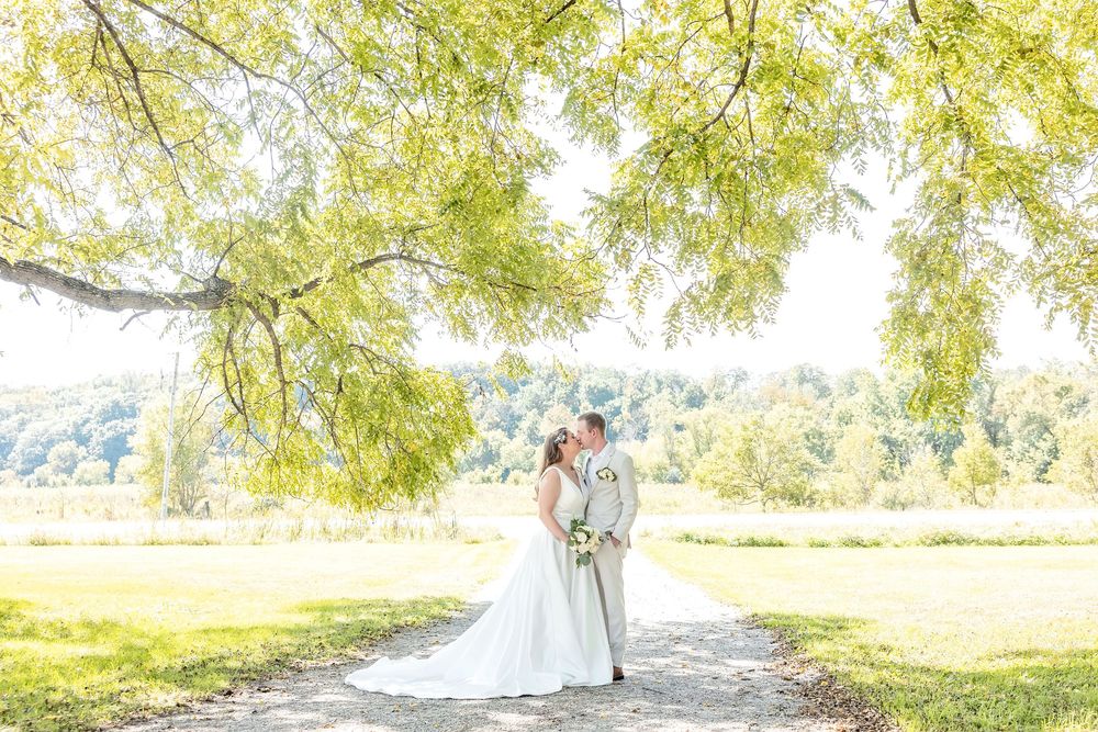 Bride and groom share a kiss under a sweeping willow tree on a sunlit garden path on their wedding day.