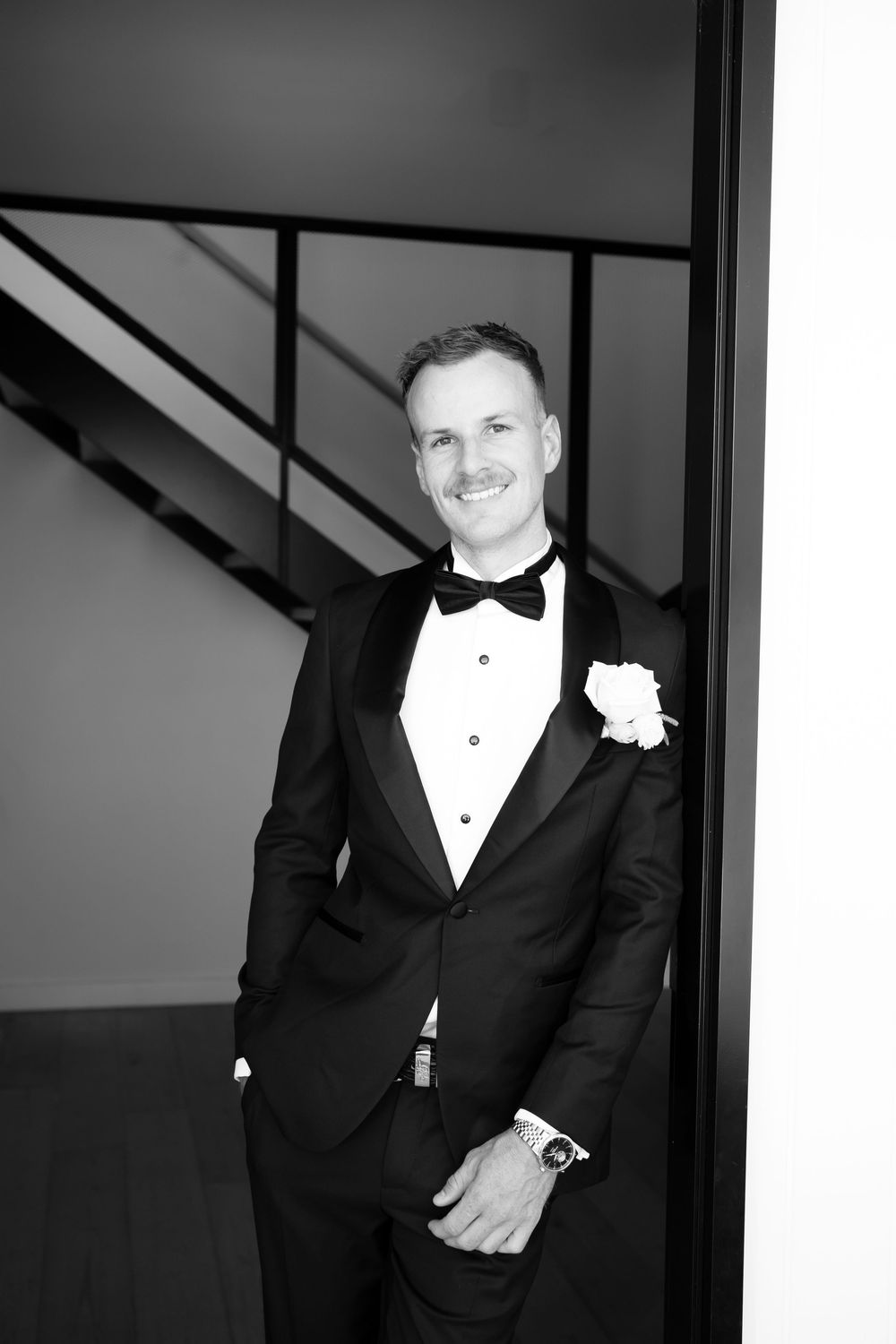 A black and white photo series showing a groom in a classic tuxedo with bow tie and boutonniere in various poses.