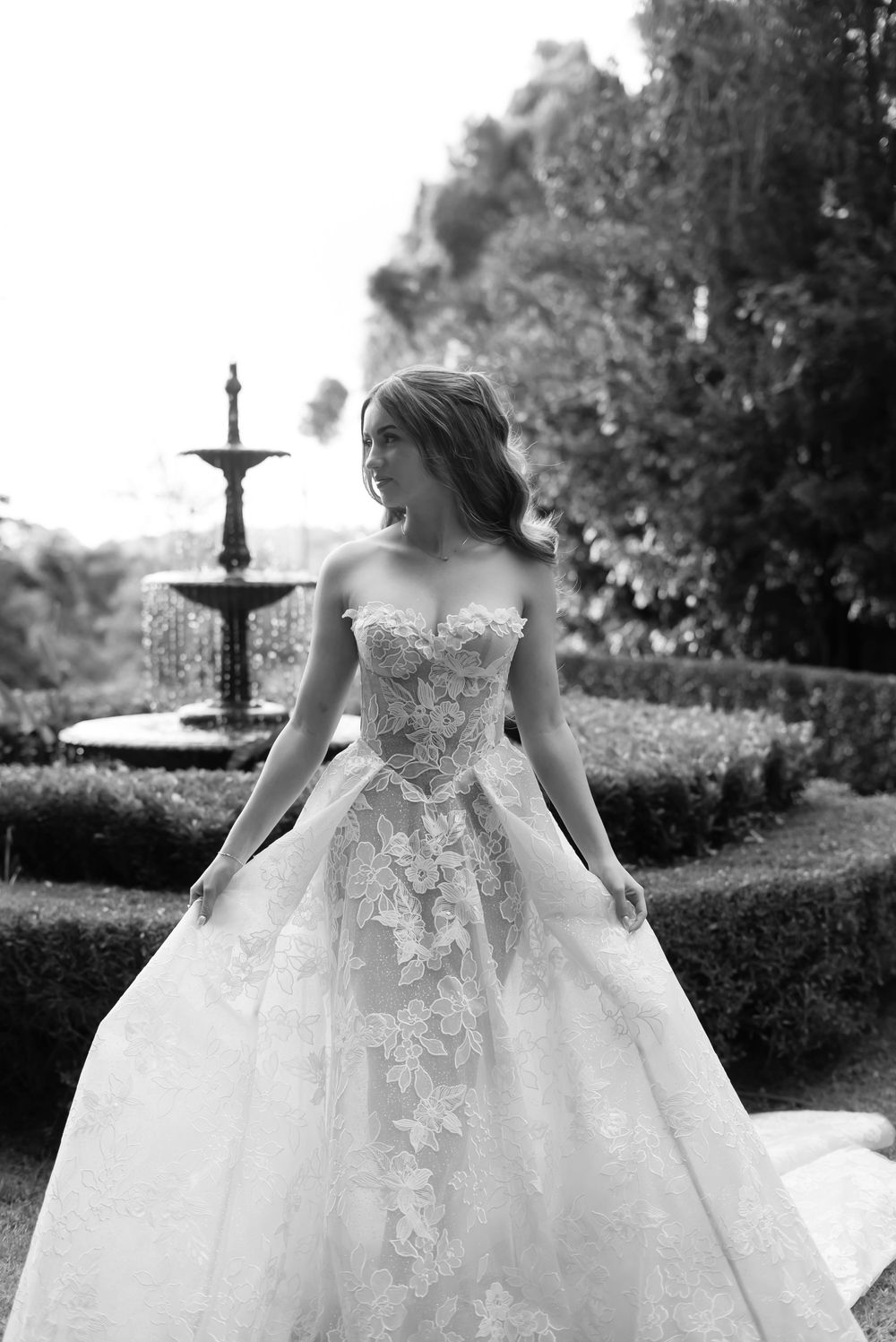 Black and white photo of an elegant wedding dress with lace details in a garden setting with a fountain in the background.