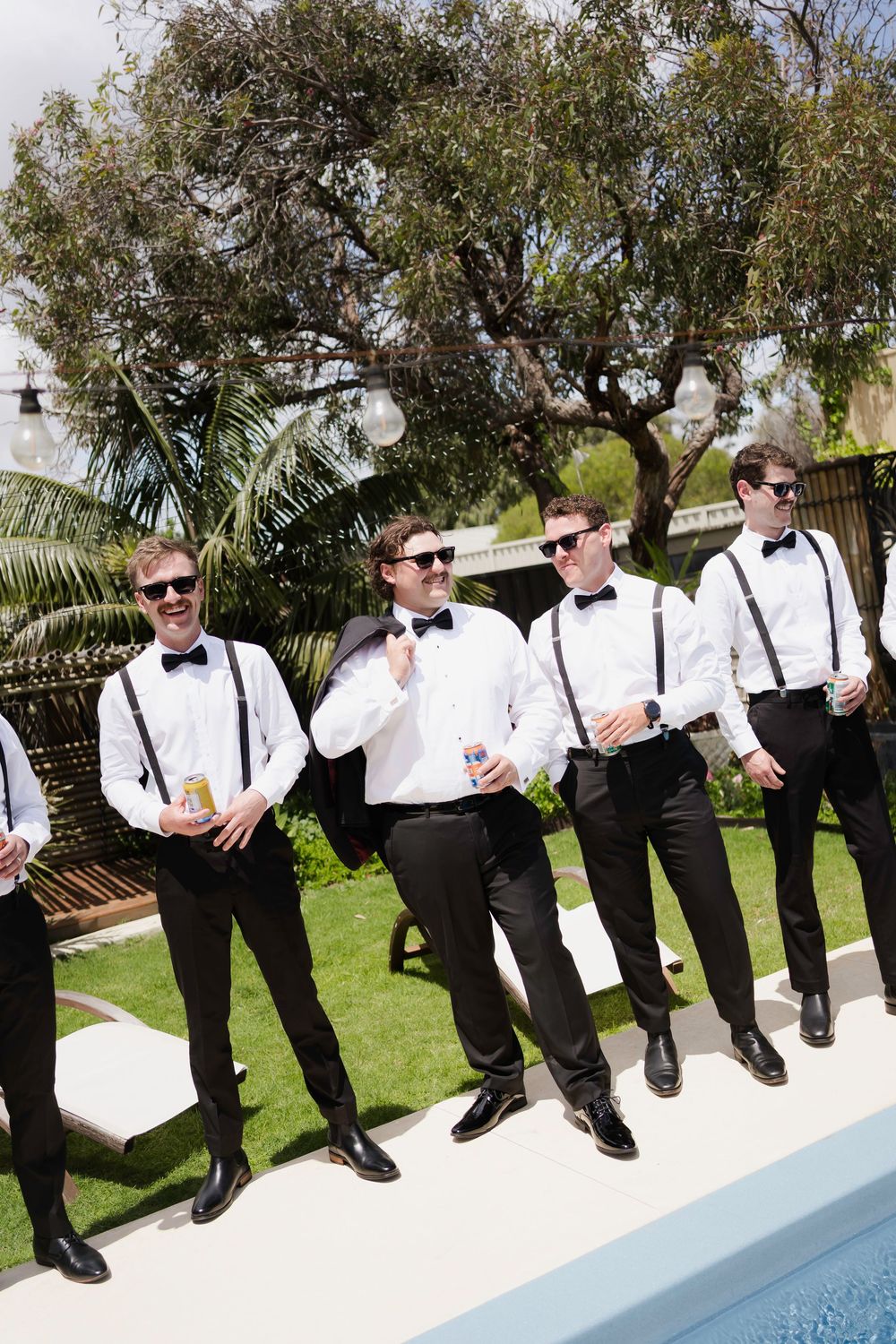 Groomsmen in black and white formal attire pose casually along a pool edge in a backyard setting.