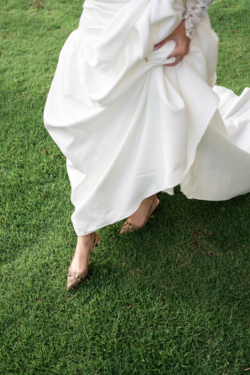 White wedding dress hemline flows gracefully over lush green grass as feet take delicate steps forward.