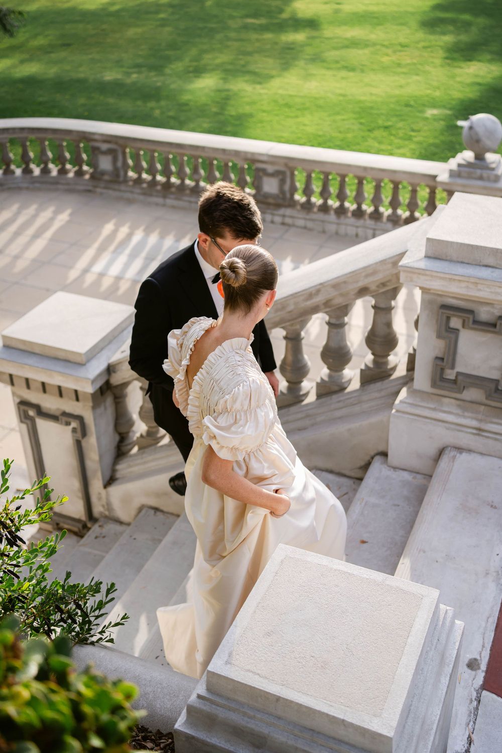 Couple in wedding attire descending ornate stone steps in afternoon sunlight.