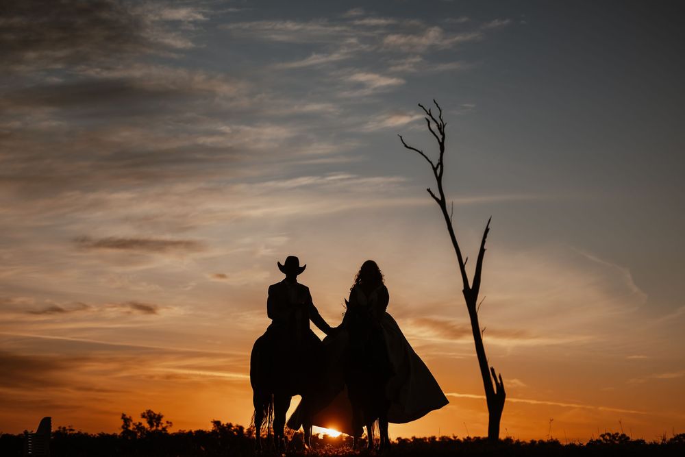 Silhouette of a cowboy on horseback and a woman in a flowing dress at sunset near a bare tree.