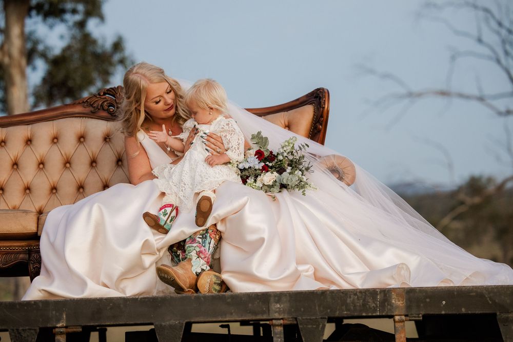 Bride in white gown sits on vintage sofa holding a baby in floral outfit, with a bouquet outdoors.