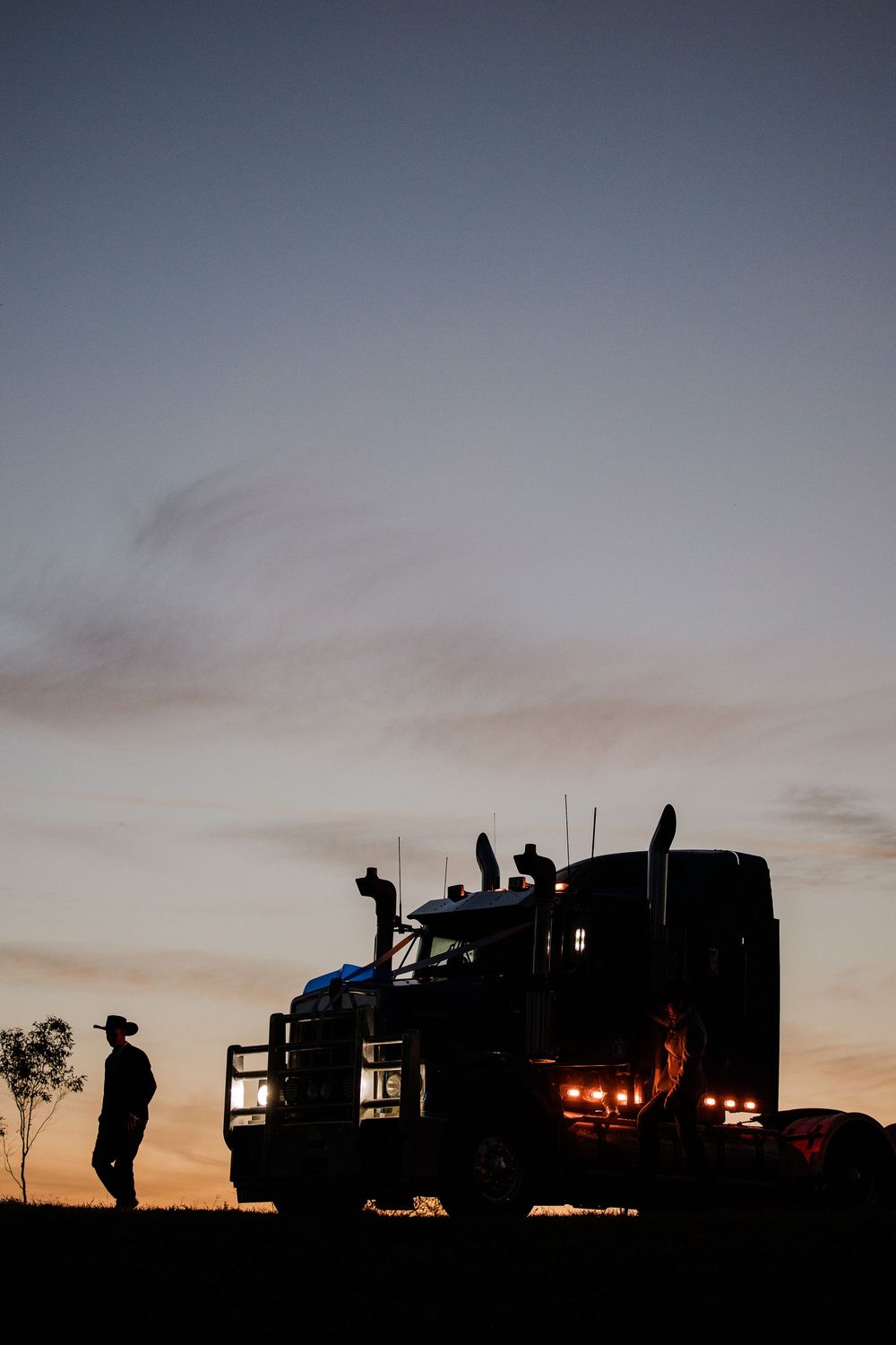 Silhouette of a truck driver standing beside a large semi-truck with glowing lights at sunset on a rural road.