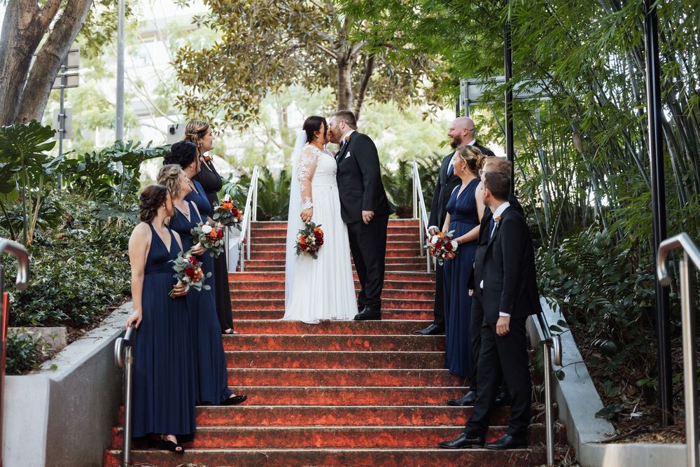 Bride and groom kiss on red stairs flanked by bridesmaids in navy dresses and groomsmen in black suits.