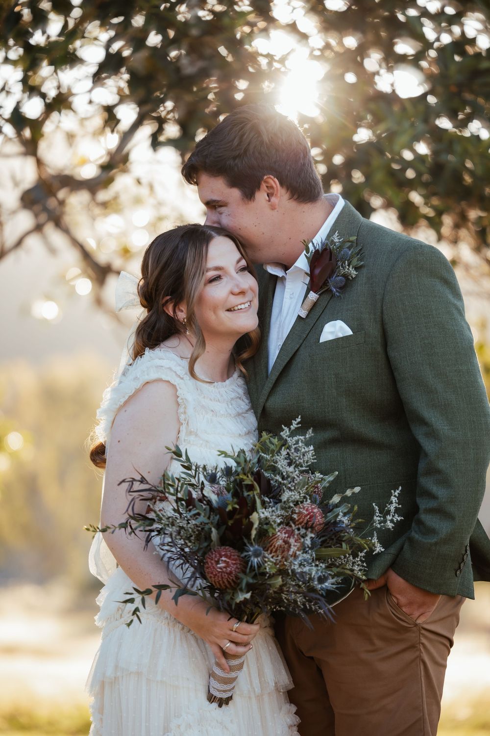 Bride and groom embrace outdoors at golden hour, bride holding a lush bouquet of proteas and greenery, groom in green tweed jacket.