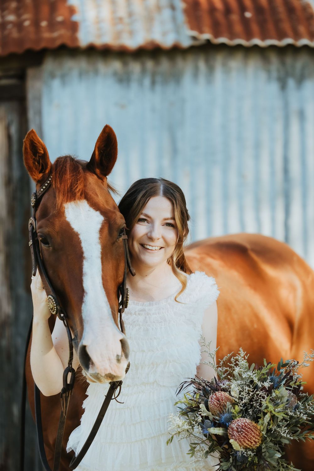 Smiling bride in white dress holding wildflower bouquet beside a chestnut horse near a rustic barn.