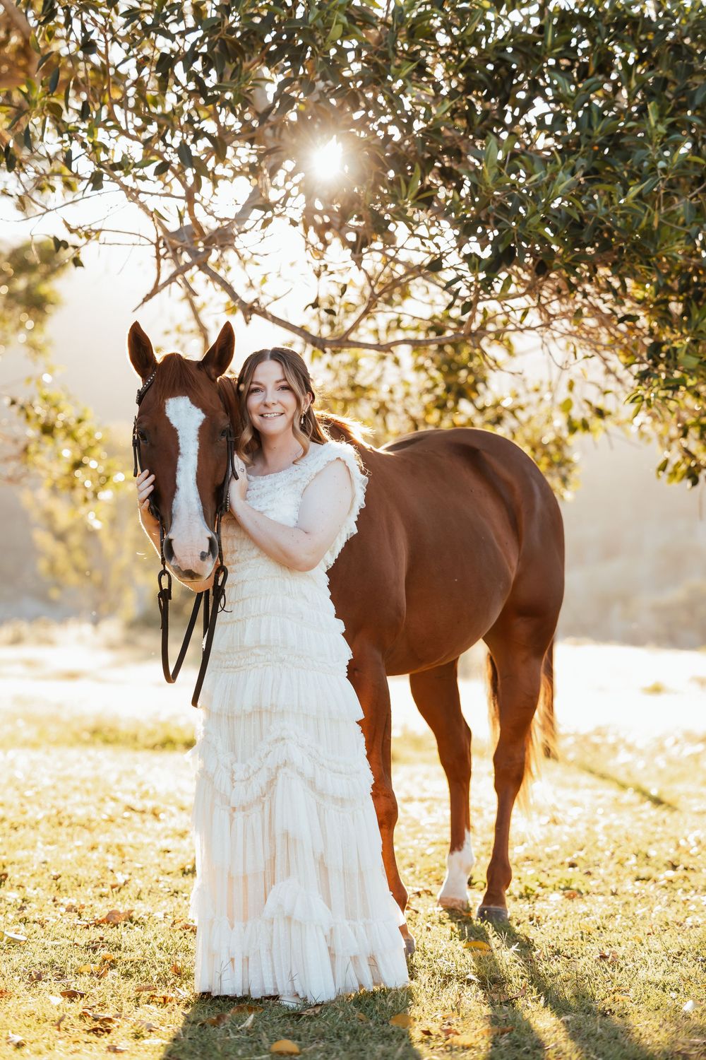 A bride in a white ruffled gown poses with a chestnut horse under sunlit trees in a golden outdoor setting.