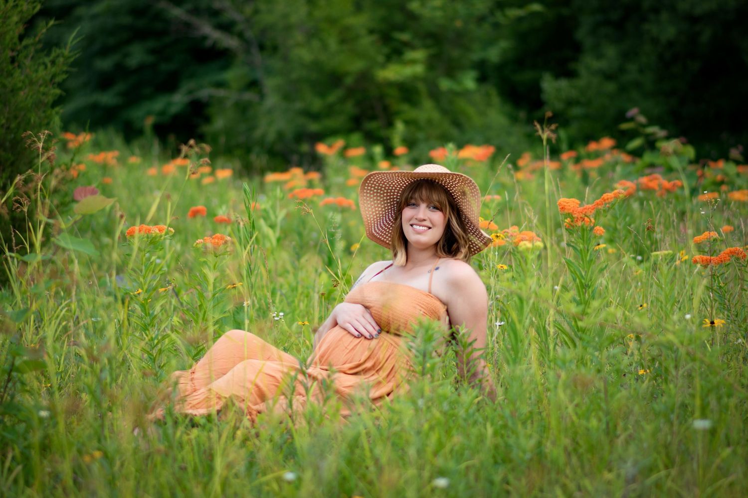 Pregnant woman in orange dress and sun hat sitting in a wildflower field smiling.