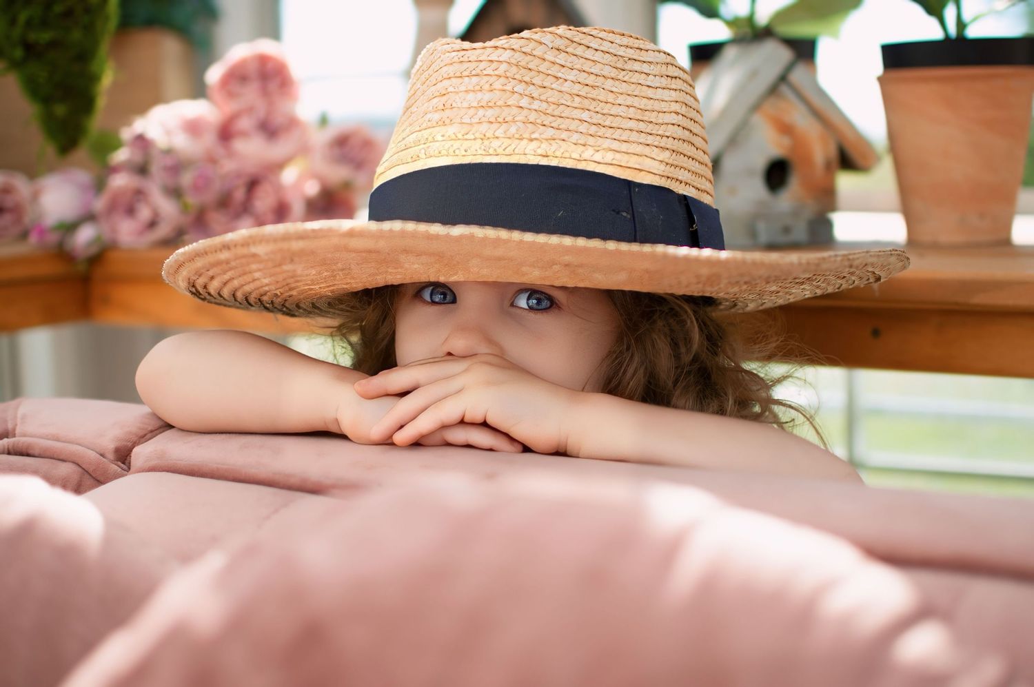 Adorable young girl peeking over a pink surface wearing an oversized straw sun hat.