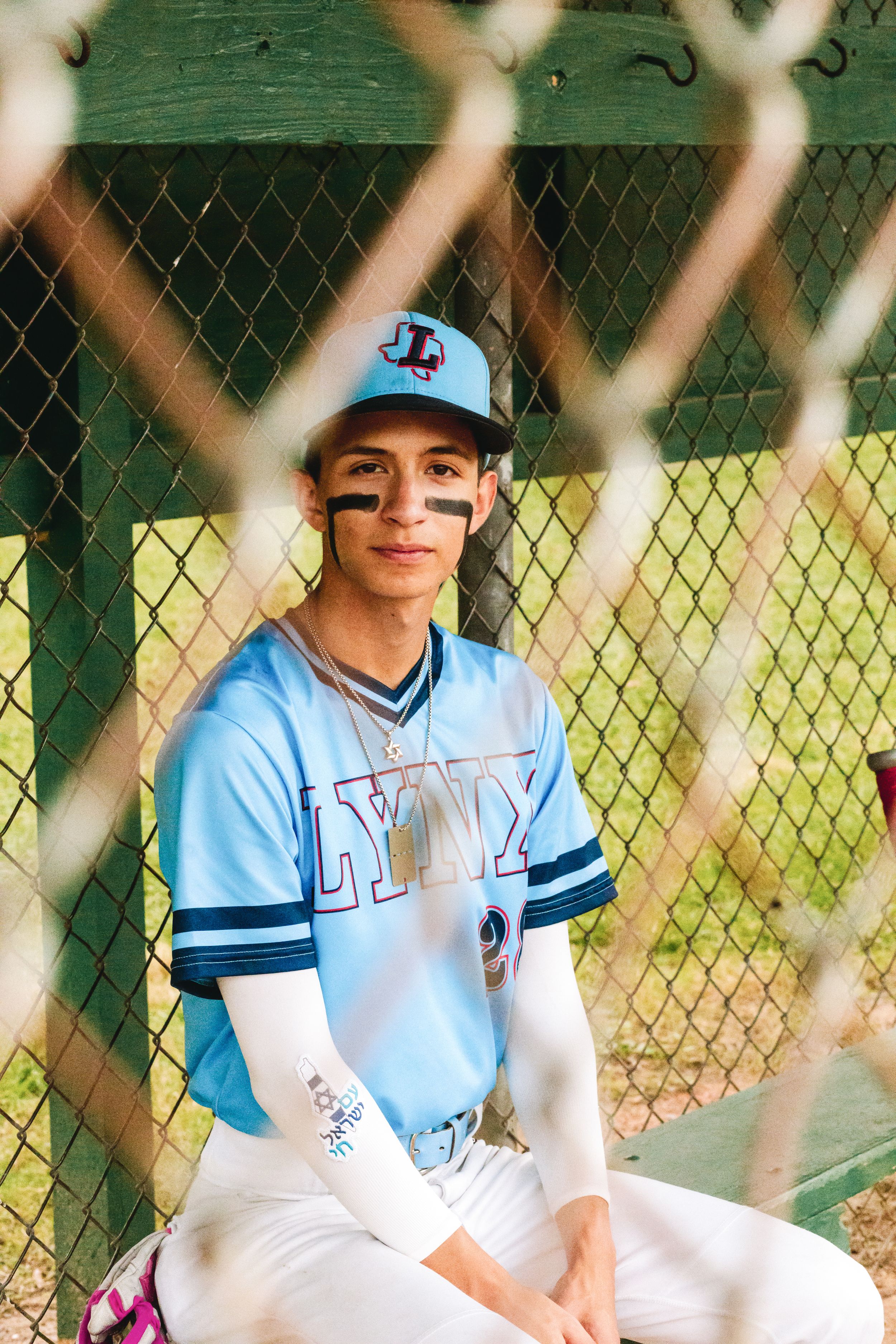 Baseball player in light blue uniform and cap sits in dugout viewed through chain link fence.