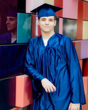 A graduate in blue cap and gown standing in front of a colorful wall with reflective panels.