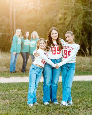 A group wearing matching varsity style sweatshirts and blue jeans pose together outdoors in a sunlit natural setting.
