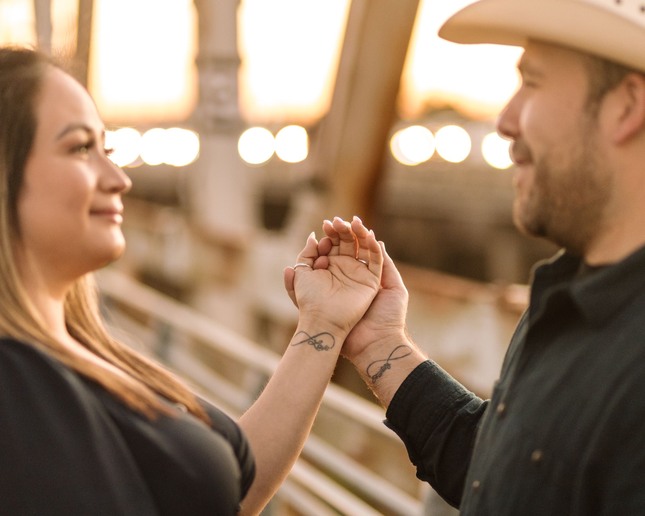 A couple holds hands and smiles at each other in warm sunset lighting with a rustic background.