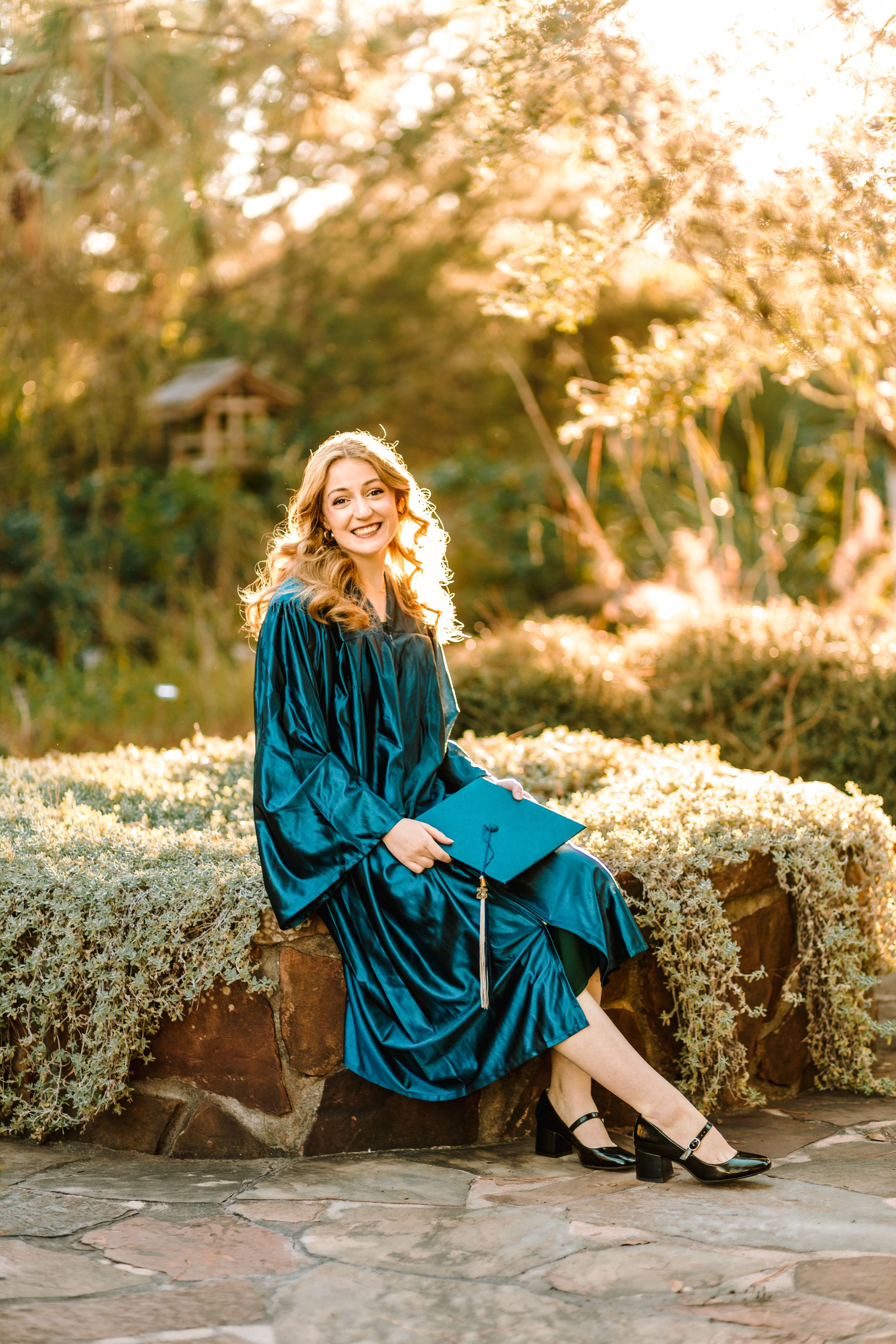 Woman in elegant teal velvet dress sits on stone wall during golden sunset hour in garden setting.
