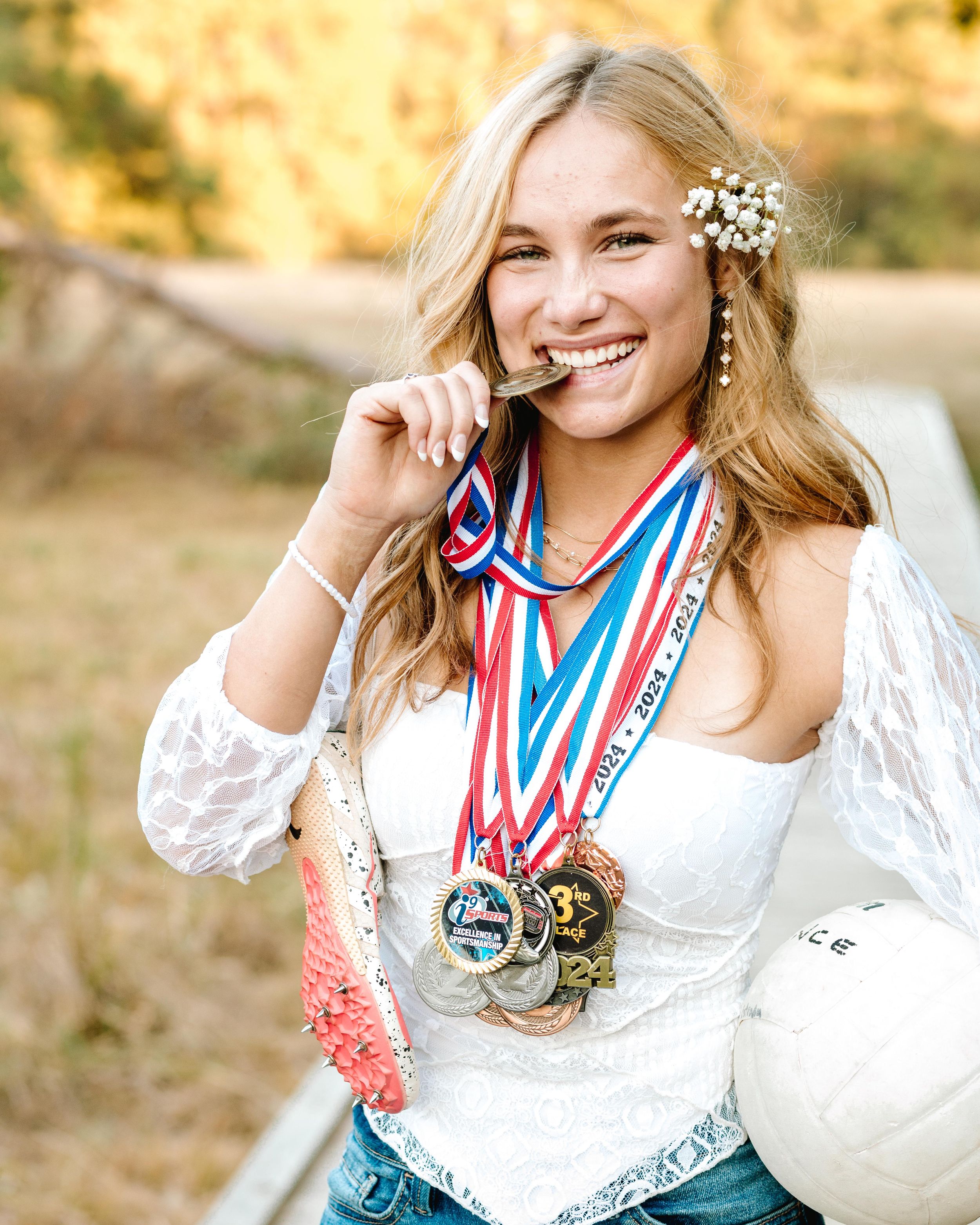 Athlete wearing white top and floral hair accessory bites medals from competitions while posing outdoors.