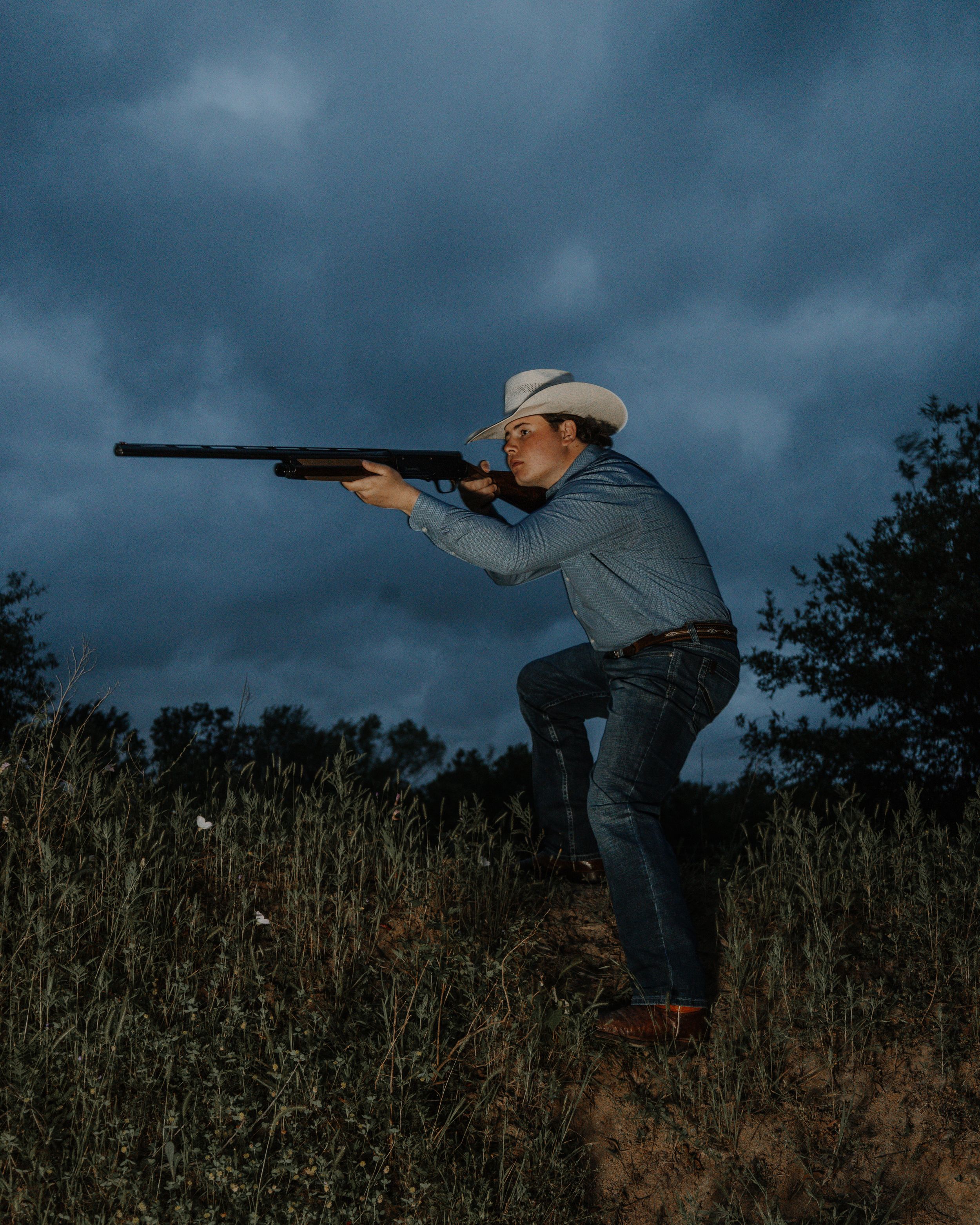 A person in jeans and a hat aims a hunting rifle while crouching on a hillside against a dramatic stormy sky.