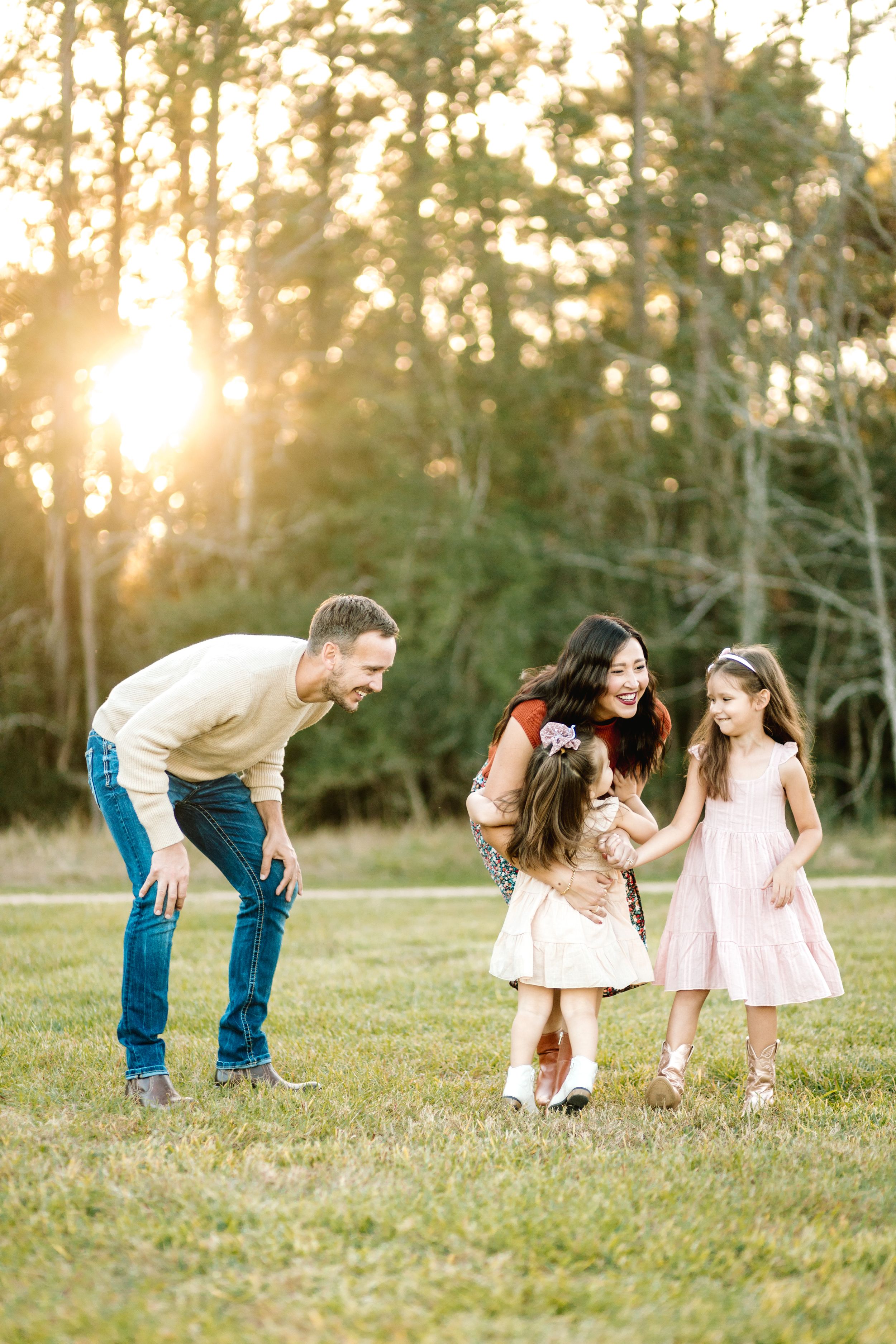 Family enjoying playful moment together in a sun-lit field at sunset with beautiful golden light filtering through trees.