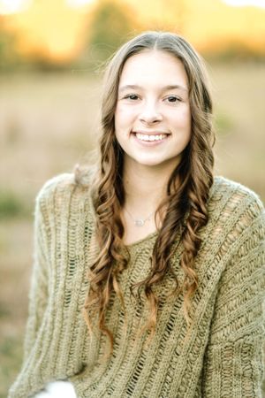 Young person with long wavy brown hair wearing a cozy knit sweater smiles against a warm autumn background.