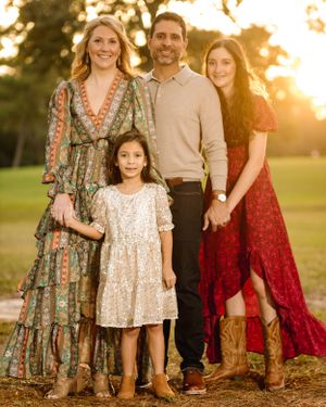 A family in coordinating outfits poses outdoors at sunset in a grassy field wearing floral dresses and casual attire.