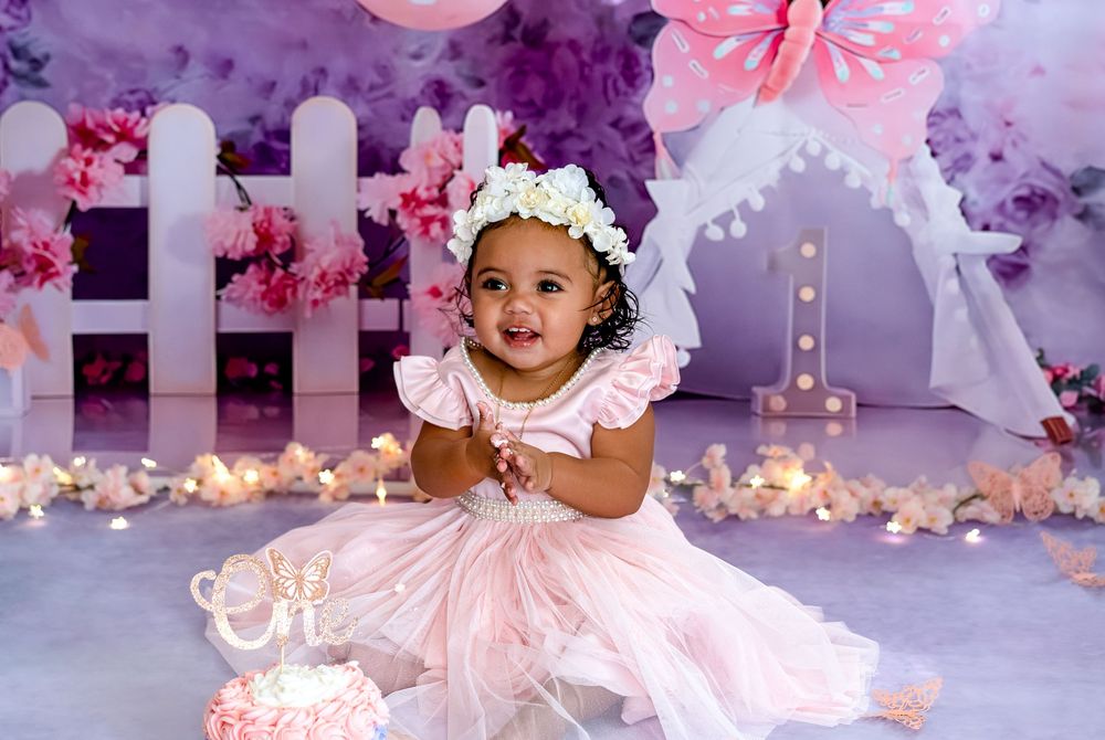 Baby in pink dress sits on floor surrounded by pink and purple decorations at a birthday celebration.