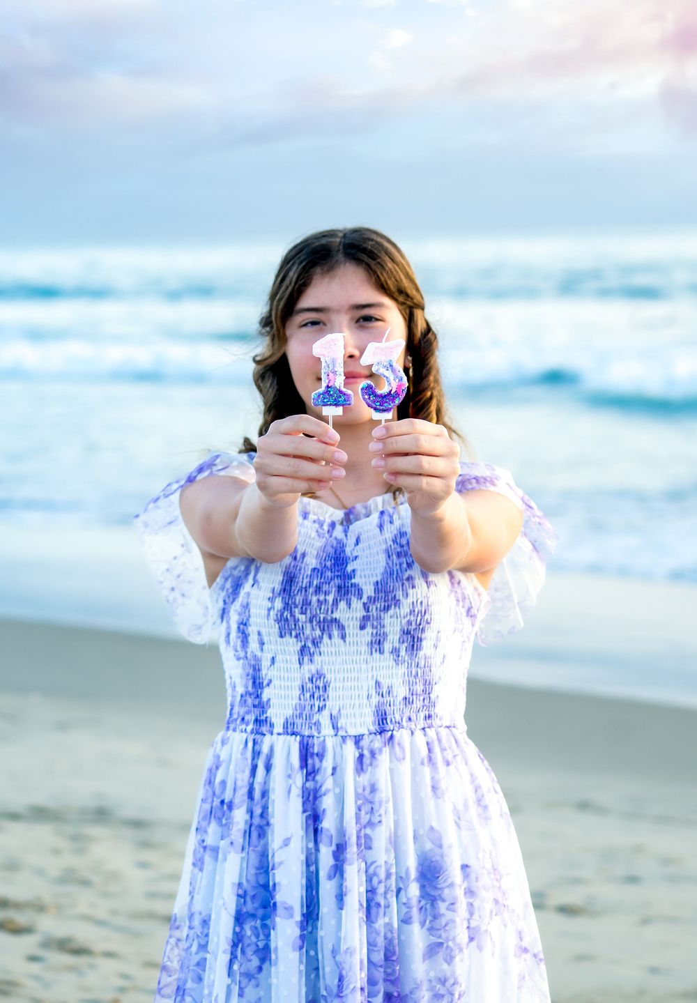 Someone in a blue and white floral dress holds up a bubble wand at the beach during sunset.