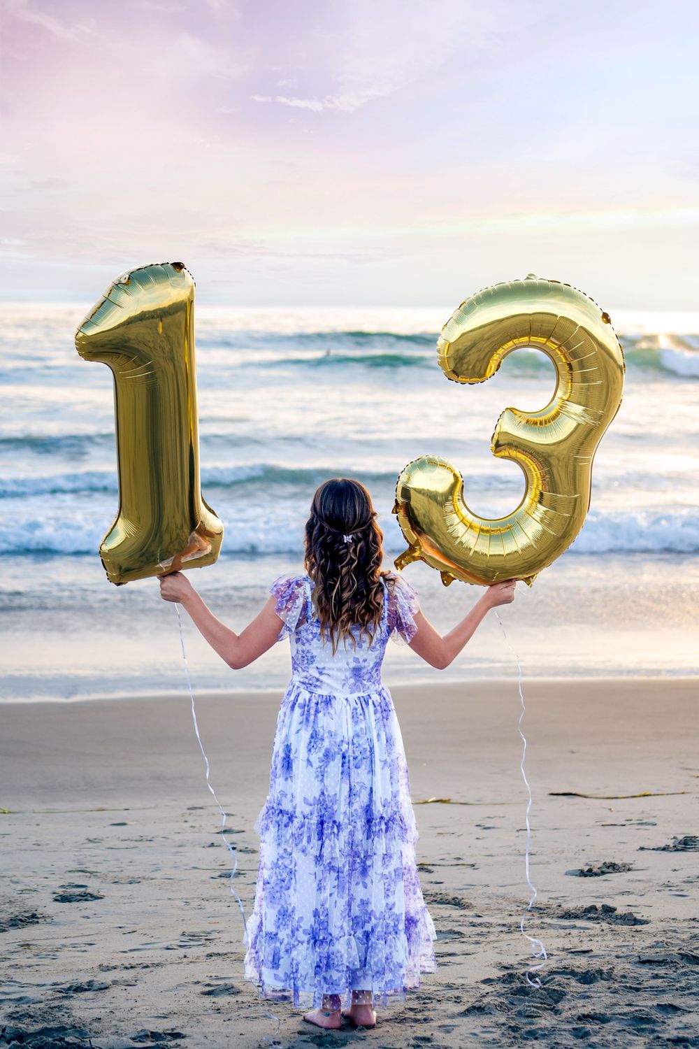 A person in a blue floral dress holds gold number balloons 1 and 3 while standing on a beach at sunset.