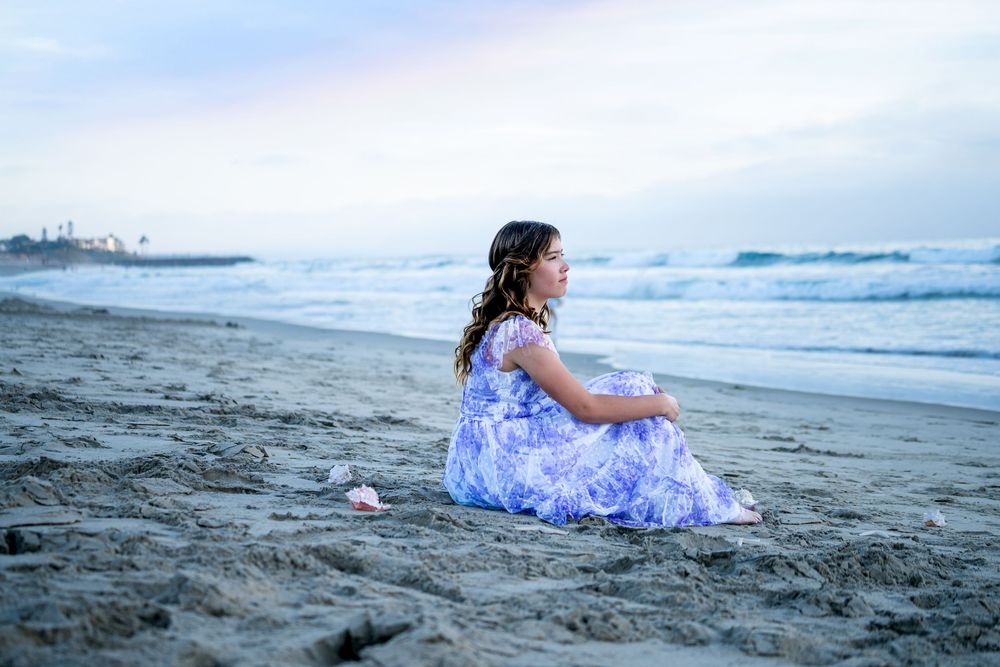 Someone in a blue and white floral dress sits alone on a misty beach at sunset watching the waves roll in.