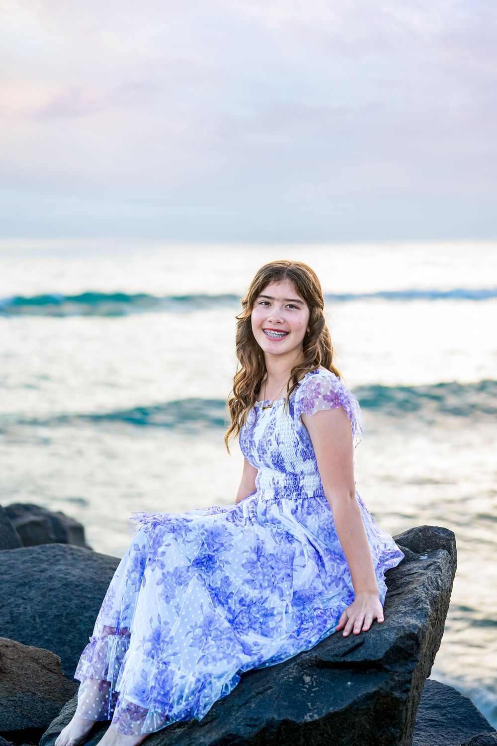 A person in a blue and white floral dress sits on dark coastal rocks at sunset by the ocean.
